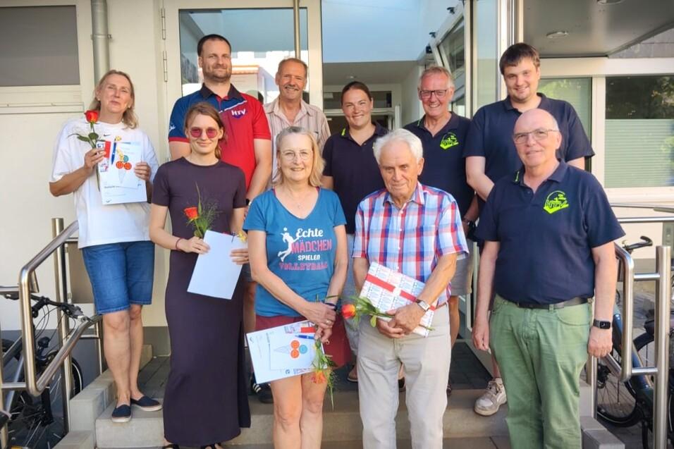 Gruppe von neun Personen vor einem Gebäude, einige halten Urkunden und Rosen, freundlich lächelnd und stehend.