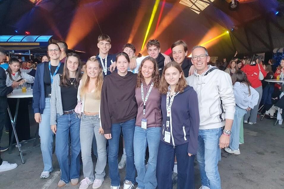 Gruppe junger Menschen posiert l&auml;chelnd in einer Veranstaltungshalle mit bunten Lichtern im Hintergrund.