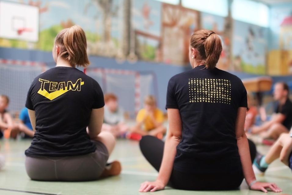 Zwei Personen mit T-Shirts, eins mit „TEAM“ und das andere mit verschiedenen Sportarten, sitzen im Kreis auf dem Boden.