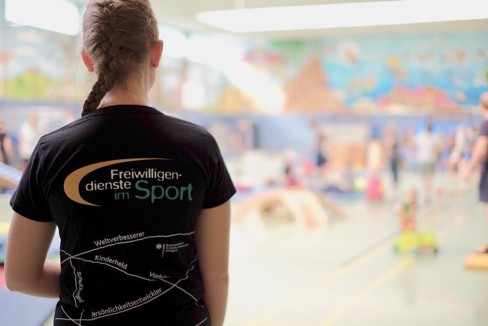 R&uuml;ckansicht einer Person mit geflochtenem Haar in einem T-Shirt mit der Aufschrift &bdquo;Freiwilligendienste im Sport&ldquo; in einer Sporthalle.