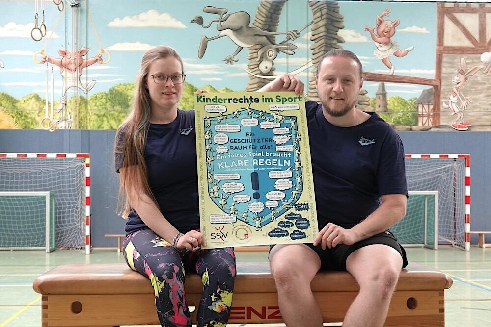 P&auml;dagogisches Duo pr&auml;sentiert ein Plakat zu Kinderrechten im Sport in einer Sporthalle mit Netz und Wandbildern.