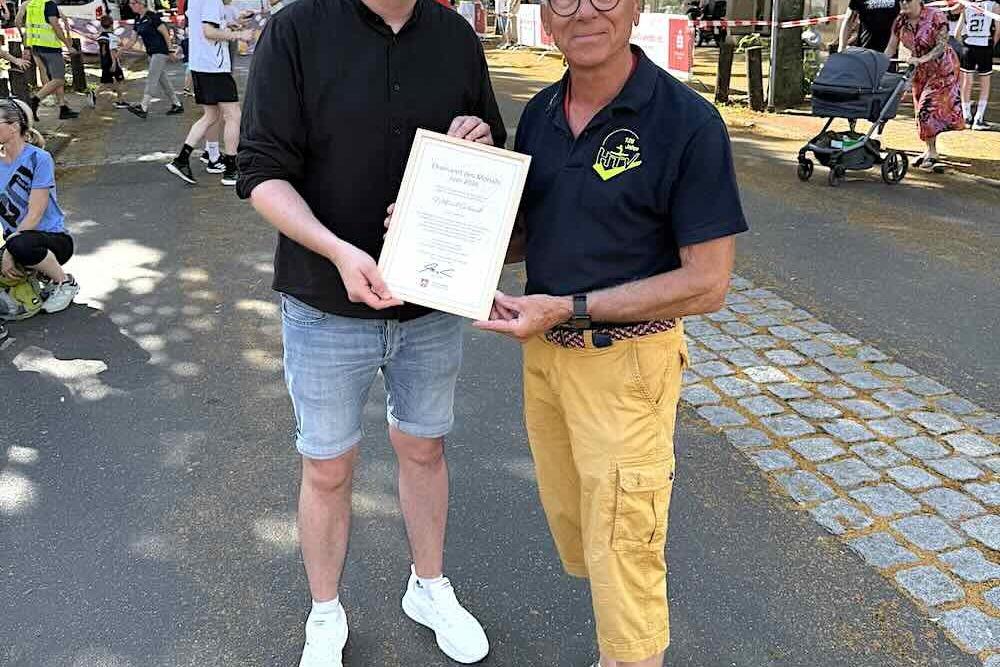 Zwei M&auml;nner stehen auf einer Stra&szlig;e, einer h&auml;lt ein Urkunde, der andere l&auml;chelt neben ihm. Im Hintergrund eine belebte Kulisse.