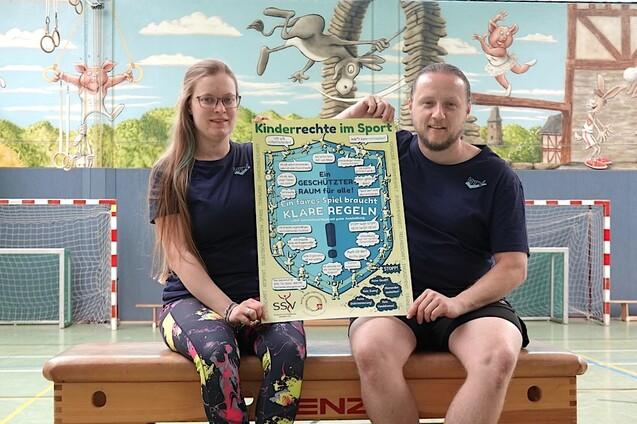 P&auml;dagogisches Duo pr&auml;sentiert ein Plakat zu Kinderrechten im Sport in einer Sporthalle mit Netz und Wandbildern.