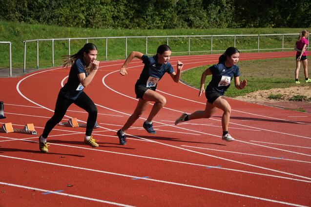 Drei junge Athletinnen starten sprintend auf einer roten Laufbahn. Sportschuhe und Startnummern sind sichtbar.