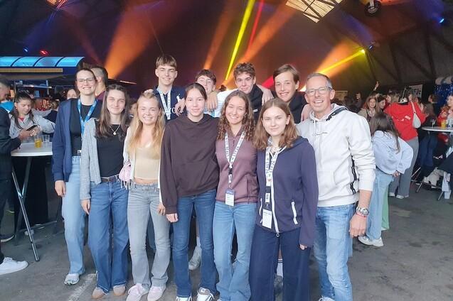 Gruppe junger Menschen posiert l&auml;chelnd in einer Veranstaltungshalle mit bunten Lichtern im Hintergrund.
