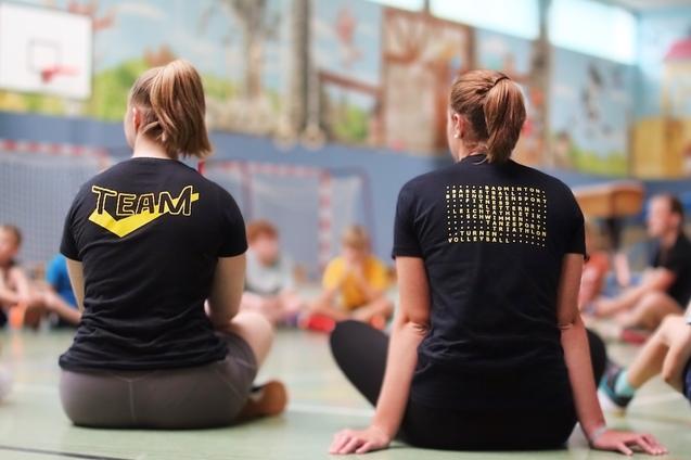 Zwei Personen mit T-Shirts, eins mit &bdquo;TEAM&ldquo; und das andere mit verschiedenen Sportarten, sitzen im Kreis auf dem Boden.