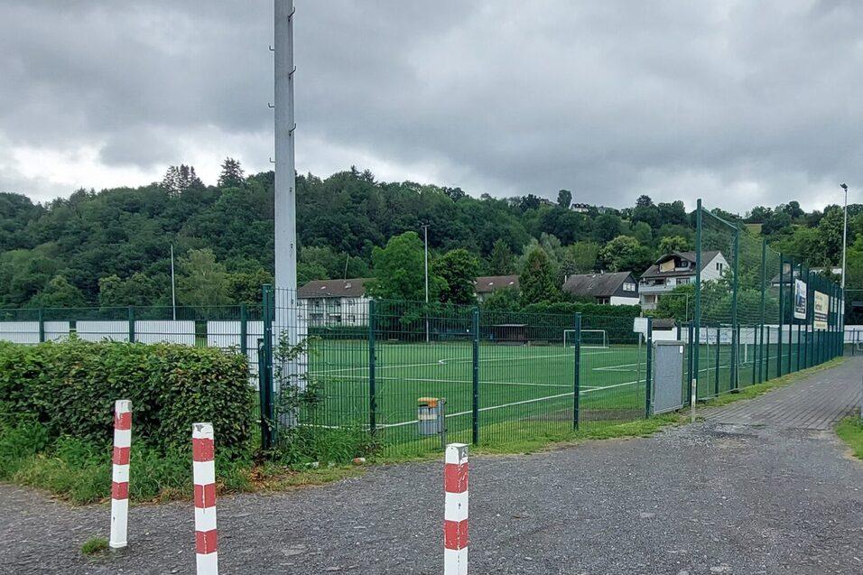 Blick auf einen Fu&szlig;ballplatz mit Zaun, umgeben von B&auml;umen und Wohnh&auml;usern unter einem bew&ouml;lkten Himmel.