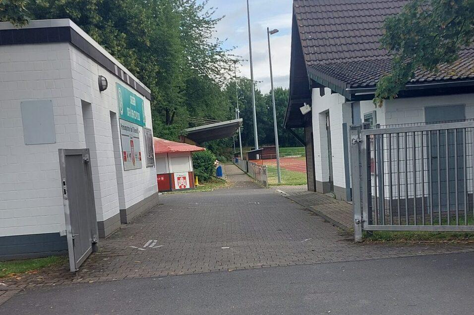 Einfahrt zu einem Sportgel&auml;nde mit zwei Geb&auml;uden und einem Blick auf den Platz im Hintergrund unter einem bew&ouml;lkten Himmel.