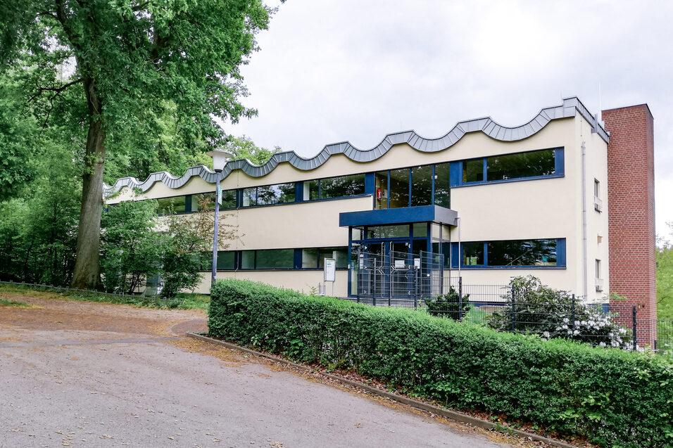 Modernes Geb&auml;ude mit wellenf&ouml;rmigem Dach, umgeben von B&auml;umen und Str&auml;uchern, an einem leicht abfallenden Weg.