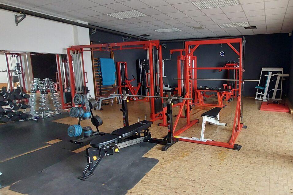 Fitnessstudio mit verschiedenen Trainingsger&auml;ten, darunter eine Langhantelbank und ein Freihantelbereich.