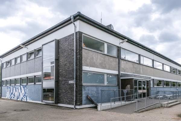 Eckansicht eines modernen, mehrst&ouml;ckigen Geb&auml;udes mit Fensterfront und grauer Fassade, umgeben von einem gepflasterten Platz.