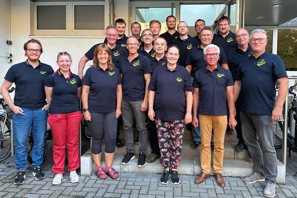 Gruppe von 20 Personen in schwarzen T-Shirts mit Logo, posierend vor einem Gebäude. Geprägt von vielfältigen Outfits und Frisuren.