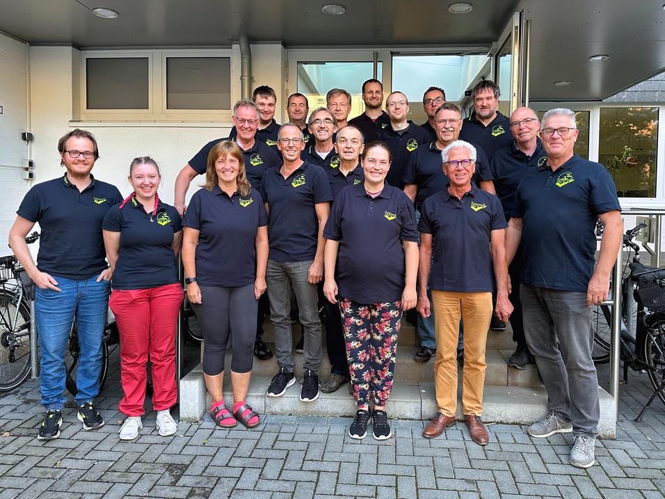 Gruppe von 20 Personen in schwarzen T-Shirts mit Logo, posierend vor einem Geb&auml;ude. Gepr&auml;gt von vielf&auml;ltigen Outfits und Frisuren.