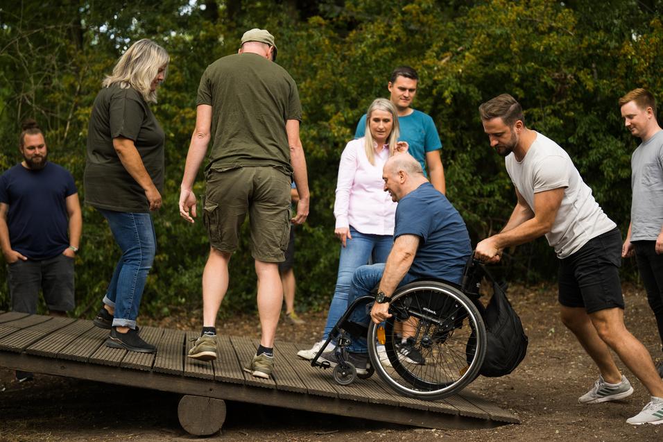 Gruppe von Menschen hilft einem Rollstuhlfahrer beim Überwinden einer Rampe in einem Waldgebiet.