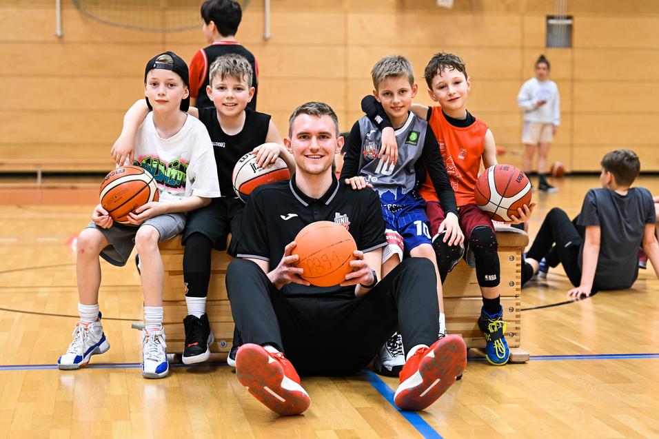 Gruppe von fünf Jungen und einem Trainer sitzt auf einer Bank in einer Basketballhalle, jeder mit einem Basketball in der Hand.