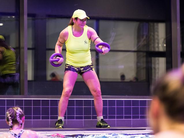 Frau in neonfarbener Sportbekleidung mit Hanteln steht am Rand eines Schwimmbeckens, im Hintergrund schwimmende Teilnehmerinnen.